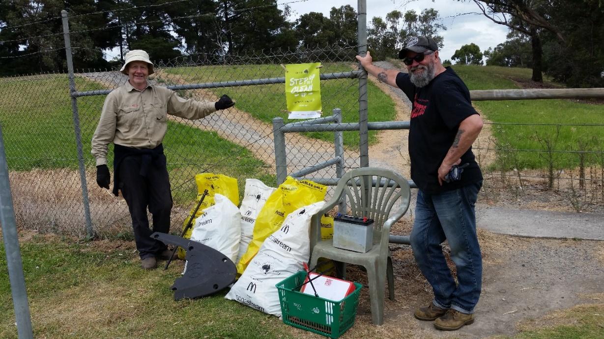 Two men standing in a grassy area with trees in the background and bags of rubbish in front of them